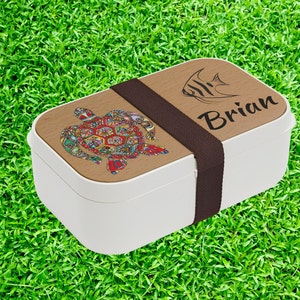 May include: White lunchbox with a brown wooden lid. The lid has a colorful turtle design, a black and white fish, and the name "Brian" written in black.