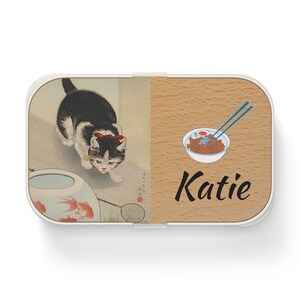 May include: A white and brown lunchbox with a Japanese-inspired design. The left side features a black and white cat with a red bow, a goldfish bowl with two goldfish, and a net. The right side has a bowl of noodles with chopsticks and the name "Katie".
