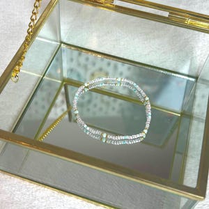 May include: A clear glass jewelry box with a gold-colored metal frame and a hinged lid. Inside, a delicate bracelet with white and iridescent beads rests on the mirrored base. A gold chain is attached to the lid.