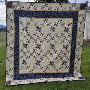 May include: A large, handmade quilt with a cream-colored background and a repeating pattern of blue and tan pinwheel shapes. The quilt has a dark blue border with a floral design and is hanging on a clothesline in a grassy area.