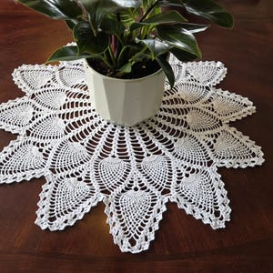 May include: A white, handmade crochet doily with a floral design, placed under a potted plant. The doily has a scalloped edge and intricate openwork patterns. The plant pot is white and the table is dark brown.