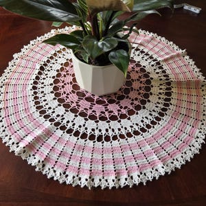 May include: A handmade, circular crochet doily in white and pink. The doily features a repeating pattern of openwork and solid stripes, with a scalloped edge. A potted plant with dark green leaves sits on top of the doily.