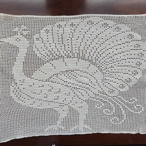 May include: Cream-colored, rectangular crocheted doily featuring a peacock design. The peacock is depicted with its tail feathers fanned out, and the entire piece is made with a delicate, openwork stitch.