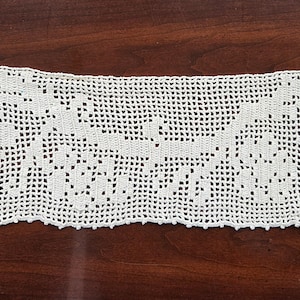 May include: Cream-colored, hand-crocheted lace trim with a floral design. The rectangular trim features openwork patterns and a scalloped edge. The lace is displayed on a dark brown wooden surface.