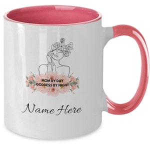Könnte beinhalten: Eine weiße Keramik-Kaffeetasse mit rosafarbenem Rand und Henkel. Die Tasse zeigt eine Strichzeichnung einer Frau mit Blumen um ihr Gesicht. Der Text "Mom by Day Goddess by Night" ist in Schwarz mit einem rosafarbenen Blumenmuster gedruckt. Der Text "Name Here" ist in Schwarz am Boden der Tasse gedruckt.