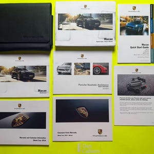 Puede incluir: Una colección de manuales y guías Porsche Macan, que incluye una funda de cuero negro, una guía de inicio rápido e información de garantía. Los manuales presentan imágenes del Macan y el logotipo de Porsche.