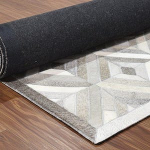 May include: A partially unrolled area rug featuring a geometric pattern composed of stitched rectangular panels in shades of grey, white, and beige. The rug has a textured hair-on-hide surface and a solid black felt backing visible on the rolled portion.