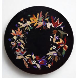 May include: A circular, black decorative plate with a floral mosaic design. The mosaic features colourful flowers in shades of red, yellow, blue, and green, arranged in a circular pattern around the edge of the plate.