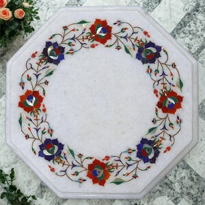 May include: Octagonal white marble table with a floral inlay. The inlay features red, blue, and green flowers and leaves in a circular pattern. The table is a decorative item, suitable for a living space.
