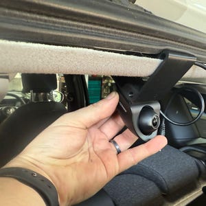 Könnte beinhalten: Eine schwarze Armaturenbrettkamera mit Weitwinkelobjektiv ist in einem Fahrzeug montiert. Die Kamera ist an einer Halterung befestigt und in der Nähe der Heckscheibe positioniert. Eine Hand hält die Kamera, und ein schwarzes Kabel ist angeschlossen.