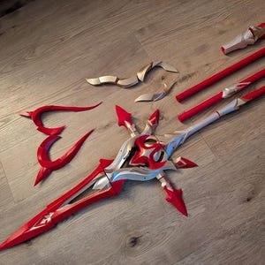 Op de afbeelding: Een rood en gouden plastic rekwisietwapen dat lijkt op een zwaard met een decoratief ontwerp. Het zwaard is in meerdere delen, waaronder een lemmet, een handvat en twee afzonderlijke delen die aan het handvat worden bevestigd.