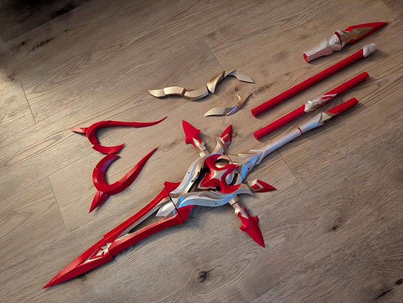 Op de afbeelding: Een rood en gouden plastic rekwisietwapen dat lijkt op een zwaard met een decoratief ontwerp. Het zwaard is in meerdere delen, waaronder een lemmet, een handvat en twee afzonderlijke delen die aan het handvat worden bevestigd.