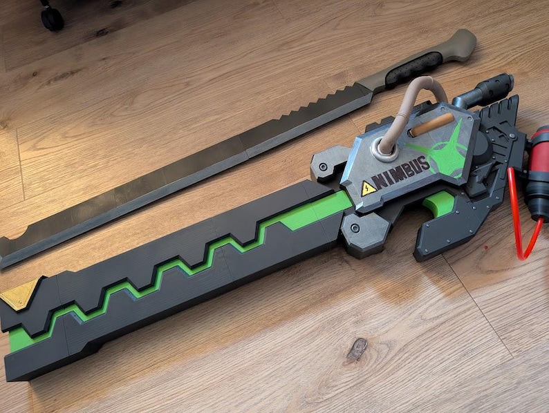 Op de afbeelding: Een zwart en groen wapen met een zilveren blad en een groen-zwarte handvat. Het handvat heeft een zilveren plaat met het woord "ANIMBUS" erop. Het wapen is een rekwisiet voor cosplay of een filmset.