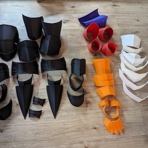 Op de afbeelding: Een verzameling 3D-geprinte kostuumstukken in zwart, wit, oranje en blauw. De stukken zijn gemaakt van plastic en zijn ontworpen om te worden gebruikt als onderdeel van een kostuum.
