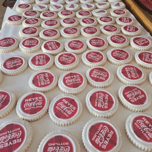 May include: A collection of round sugar cookies with white icing and a red and white Coca-Cola bottle cap design. The cookies are arranged on a white surface, with the text "REYES Coca-Cola BOTTLING" visible on each cookie.