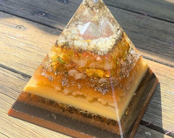 May include: A layered pyramid sculpture with a clear resin exterior. The pyramid features alternating bands of brown, black, and yellow, with embedded gold flakes and crystals. The top of the pyramid has a white crystal. The pyramid is sitting on a wooden surface.