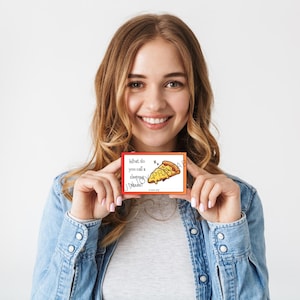 K&ouml;nnte beinhalten: Eine kleine, rechteckige Karte mit rotem Rand und dem Text "What do you call a sleeping pizza?" zeigt ein Cartoon-Pizzast&uuml;ck mit geschlossenen Augen. Die Karte wird von einer Person gehalten, die eine Jeansjacke tr&auml;gt.