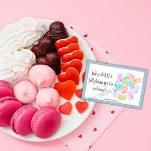 K&ouml;nnte beinhalten: Ein wei&szlig;er Teller mit rosa Makronen, herzf&ouml;rmigen S&uuml;&szlig;igkeiten und rosa Baisers. Auf der rechten Seite befindet sich eine Karte mit dem Text "Why did the jellybean go to school?" vor rosa Hintergrund. Valentinstag-Thema.