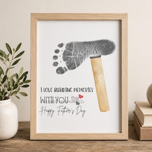 May include: Framed print with a gray footprint and a hammer illustration. The text reads "I LOVE BUILDING MEMORIES WITH YOU" and "Happy Father's Day". The frame is light brown.