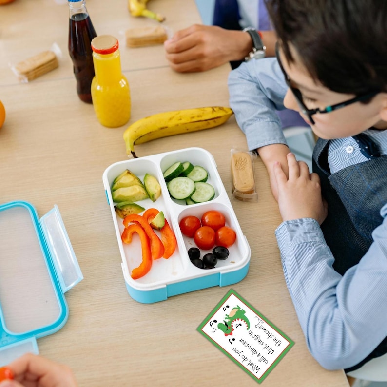 K&ouml;nnte beinhalten: Eine geteilte, wei&szlig;-blaue Lunchbox mit Avocadoscheiben, Gurkenscheiben, Paprikastreifen, Kirschtomaten und schwarzen Oliven. Eine gelbe Banane und eine Saftflasche sind in der N&auml;he. Eine Karte mit einer gr&uuml;nen Dinosaurier-Illustration und Text ist ebenfalls vorhanden.