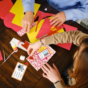 K&ouml;nnte beinhalten: H&auml;nde basteln Valentinstagskarten mit rotem, gelbem und rosa Papier, Schere und Kleber. Eine Karte zeigt eine Computergrafik und den Text "Happy Valentine's Day". Eine andere Karte hat eine Astronautengrafik und den Text "Why did the astronaut bring a pencil to space?"