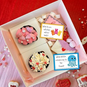 K&ouml;nnte beinhalten: Eine Geschenkbox zum Valentinstag mit Leckereien. Enth&auml;lt zwei Cupcakes mit rosa Zuckerguss und Schmetterlingsdekorationen, Kekse und Karten mit Witzen. Die Box ist wei&szlig; mit rosa Innenfutter.