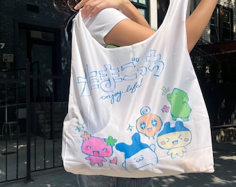 Könnte beinhalten: Weiße wiederverwendbare Tragetasche mit einem japanischen Spruch und Comicfiguren. Der Spruch lautet "Genieße das Leben!" und die Figuren sind ein rosa Wesen, ein blaues Wesen, ein gelbes Wesen mit einem Wirbel auf dem Kopf und ein grüner Frosch.