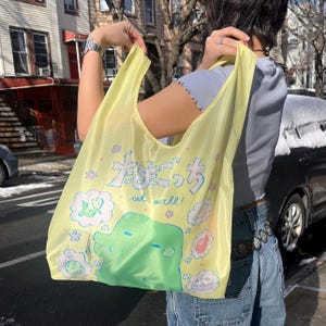 Puede incluir: Una bolsa de compras reutilizable amarilla con una ilustración de dibujos animados de una criatura verde y el texto "Eat well!" en inglés y japonés. La bolsa tiene una correa blanca y está hecha de un material ligero.