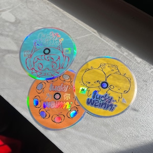 May include: Three round holographic stickers with cartoon characters and text. The first sticker is blue with a cartoon character wearing a red hat and the text "Lucky Weini" in a bubble. The second sticker is orange with a cartoon character holding a red apple and the text "Lucky Weini" in a bubble. The third sticker is yellow with two cartoon characters and the text "Lucky Weini" in a bubble.