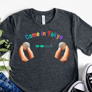 May include: Dark grey t-shirt with the text "Come in Tokyo" in rainbow colours. The text is surrounded by two hands holding a silver coin.