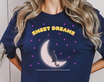 Op de afbeelding: Een marineblauw t-shirt met de tekst "Sweet Dreams" in gele en roze letters. Een zwart katten silhouet zit op een halve maan met roze sterren op de achtergrond.
