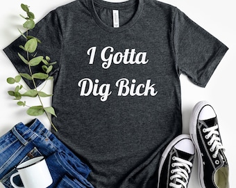 May include: A dark grey t-shirt with the text "I Gotta Dig Bick" in white lettering. The shirt is laid flat on a white surface with a pair of black canvas shoes and a pair of blue jeans.