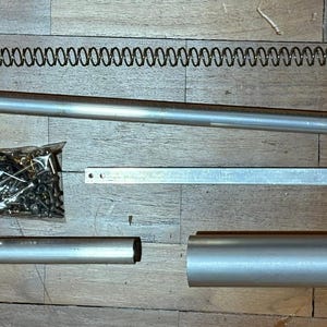 Puede incluir: Un surtido de componentes de hardware metálicos, que incluyen un resorte largo, tubos cilíndricos, una barra rectangular y una pequeña bolsa de tornillos y otras piezas pequeñas. Los artículos están dispuestos sobre una superficie de madera.