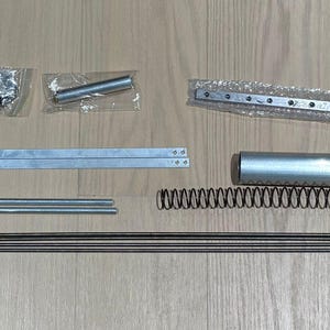 May include: A collection of metal hardware components, including rods, springs, and brackets, arranged on a wooden surface. The items are in various shapes and sizes, with silver and black finishes. The components appear to be for a mechanical assembly.