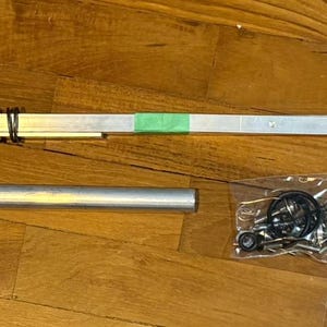 Pode incluir: Uma variedade de componentes metálicos, incluindo uma barra longa e fina de prata com uma mola e fita verde, um tubo cilíndrico e um pequeno saco de hardware. Os itens estão dispostos em uma superfície de madeira.
