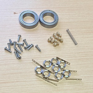 May include: A collection of mechanical components, including two ball bearings, screws, nuts, springs, and retaining rings. The parts are made of metal and arranged on a light-coloured surface.