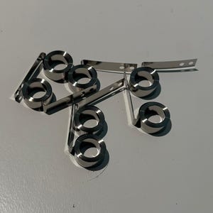 Pode incluir: Uma coleção de componentes metálicos prateados e brilhantes. A imagem mostra vários anéis circulares com uma pequena tira de metal plana anexada. As peças estão dispostas sobre uma superfície clara, projetando sombras.