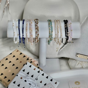 May include: A set of eight beaded bracelets in various colours, including white, blue, pink, brown, and black, displayed on a white stand. The bracelets are made of small, round beads and have a simple, elegant design. The bracelets are packaged in gift boxes with a "A Gift for You" tag.
