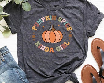 May include: Dark grey heather t-shirt with a circular graphic that reads "Pumpkin Spice Kinda Girl" around an orange pumpkin illustration. The image also includes blue jeans and brown sandals.
