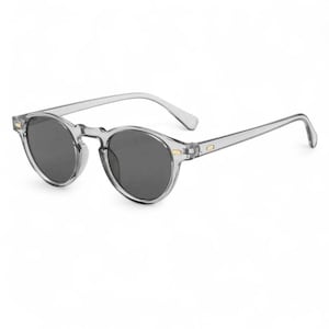 Peut inclure: Lunettes de soleil rondes à monture transparente avec verres gris foncé. Les montures et les branches sont d'un gris transparent, avec de petits accents dorés au niveau des charnières. Style rétro classique.