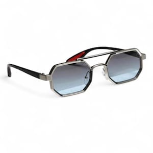Puede incluir: Gafas de sol con montura plateada y forma octogonal. Las lentes presentan un degradado de gris oscuro a azul claro. Las patillas son negras con un detalle rojo. Un doble puente completa el diseño.