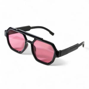 May include: Black sunglasses featuring a geometric frame and pink lenses. The sunglasses have a double bridge and a modern design. The lenses are a vibrant pink, contrasting with the black frame. A stylish accessory.