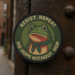 May include: Circular patch featuring a green frog illustration with a blue scarf. The patch has the words "RESIST. REPEAT" at the top and "NO HOPE WITHOUT HOP" at the bottom, in white text. The frog is set against a green backdrop.