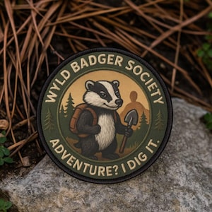 Könnte beinhalten: Runder Aufnäher mit dem Text "WYLD BADGER SOCIETY" und "ADVENTURE? I DIG IT." Er zeigt einen Dachs mit Rucksack und Schaufel vor einer Waldszene mit beigem Hintergrund und grünem Rand.