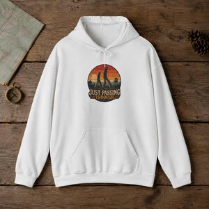 May include: A white hooded sweatshirt featuring a circular graphic on the chest. The design displays a sunset background with two silhouettes of Bigfoot walking and the text "JUST PASSING THROUGH" in a bold, dark font centered below the figures.