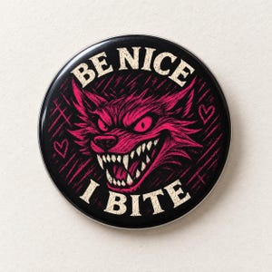 May include: A circular pin-back button featuring a black background with a stylized, magenta-colored wolf head graphic. The wolf has bared teeth and glowing eyes. The text BE NICE I BITE is printed in a cream-colored font, accented by small hearts.
