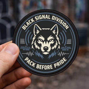 May include: A circular black patch featuring a wolf's head at the centre, with the text "BLACK SIGNAL DIVISION" and "PACK BEFORE PRIDE" around the edge. The wolf is white and grey, with a detailed design. The patch has blue and grey accents.
