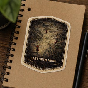 May include: A sticker with a vintage map design featuring red push pins marking locations. The sticker has a distressed border and the text "LAST SEEN HERE." is printed at the bottom. The sticker is on a spiral notebook.