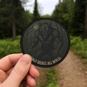 May include: A round patch featuring a werewolf illustration in shades of gray and black, set against a forest backdrop with a full moon. The text "HALF BEAST. ALL WYLD." is printed around the bottom edge.
