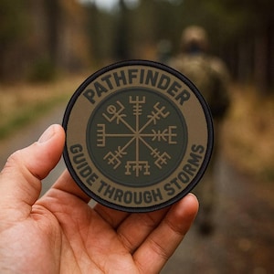 Pode incluir: Remendo redondo com o texto "PATHFINDER GUIDE THROUGH STORMS". O remendo apresenta um desenho de rosa dos ventos no centro, com uma borda castanha e uma borda preta. O remendo é segurado em uma mão.
